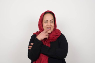 Hayalperest Müslüman kadın, düşünceli bir ifadeyle, kameraya bakıyor, yüzüne yakın duruyor, parmağını ısırıyor, hoş bir şey düşünüyor, gri duvara karşı poz veriyor. Stüdyoda poz veren başörtüsü takan orta yaşlı bir Arap kadın.