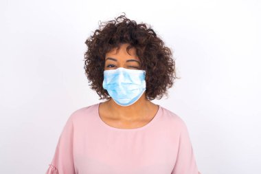 Yüz maskesi takan ve göz kırpan kadın beyaz stüdyo arka planında izole edilmiş.