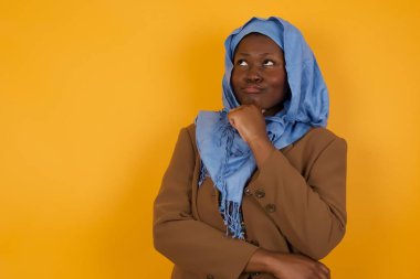 Düşünceli gülümseyen Afrikalı Amerikalı Müslüman kadının portresi elini çenesinin altında tutuyor, yan bakıyor, ilgi çekici bir şey düşünüyor ya da merak ediyor sarı stüdyoya karşı gelişigüzel poz veriyor. Karar alma kavramı. 