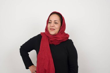 Neşeli olgun Müslüman kadının stüdyo çekimi elini kalçasında tutuyor, geniş bir gülümseme sergiliyor, arkadaşlarının doğum gününde hazırladığı sürprizi bekliyor, arka planda izole edilmiş bir şekilde..