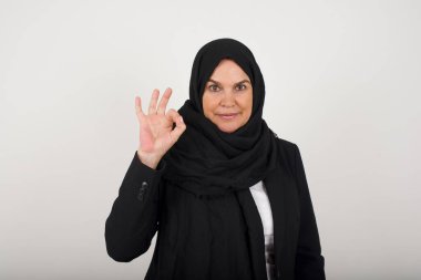 Siyah tesettürlü çekici müslüman kadının, onaylandığı gibi işaret diliyle işaret vermesine, neşeli bir ifadeye sahip olmasına sevindim. Güzel bir kadının fotoğrafı çekici bir görünüme sahip, iyimser olmak. Gri duvara yaslanarak.