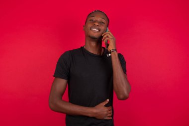 Kırmızı arka planda siyah tişört giyen ve cep telefonuyla konuşan gülümseyen, sıradan, başarılı bir Afrikalı Amerikalı 'nın portresi. İş, güven ve iletişim kavramı.