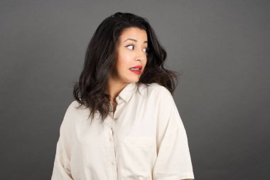 Güzel esmer kadın, sıradan giyinmiş, gözlerini kaçırıyor. Gri arkaplanda duruyor.  