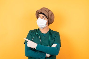 Yeşil üniformalı güzel, genç, Müslüman doktor kadın kameraya hileli bir şekilde poz veriyor ve bir yandan el kol hareketi yapıyor. Bayan tasarımcı, işaret parmağıyla harika bir ürün sunuyor. Hareket kavramı.