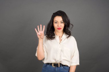 Gayri resmi giyinmiş güzel esmer kadın dört parmağını gösteriyor. Gri arkaplanda duruyor. 
