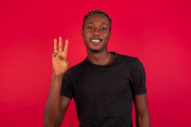 Genç sarışın adam duvarın önünde duruyor. Üç numaralı parmaklarıyla işaret ediyor ve gülümsüyor. Kendinden emin ve mutlu..