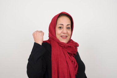 Sevinçten havalara uçmuş mutlu, mutlu bir kadın, iyi haberler aldığına sevinir, mutluluktan hareketler yapar, yumruğunu sıkar ve kazanan jestler yapar. Tepki konsepti. Stüdyoda poz veren başörtüsü takan orta yaşlı bir Arap kadın.