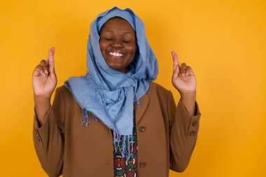 Kazanmak zorundayım. Neşeli genç Afrikalı Afrikalı Amerikalı Müslüman kadın dişlerini sıkıyor, parmaklarını çapraz yapıyor, arzulanan dilekler diliyor, gündelik kıyafetler giyiyor, iyi haberleri bekliyor, sarı duvarın üzerinde duruyor.