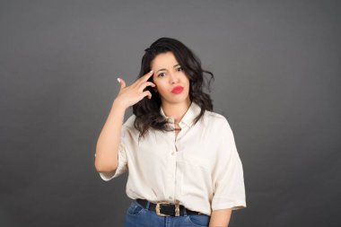 Güzel esmer kadın kayıtsız şartsız giyinmiş. Gri arka planda silah hareketi yapıyor..  