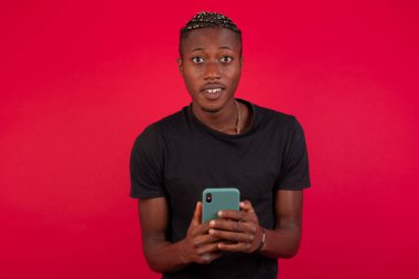 Kırmızı arka planda siyah t-shirt giyen Afro-Amerikalı bir adam kameraya odaklan, akıllı telefondan uzak dur, gülümse ve mutlu bir şekilde cep telefonunu tut, aynada selfie çek, arkadaşlarına yeni kıyafetlerini göster