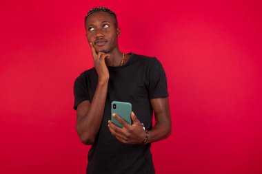 Düşünen genç bir Afro-Amerikalı adamın kırmızı arka plan üzerinde siyah tişört giyip cep telefonu kullandığını ve el ele tutuştuğunu hayal et. Karar almak ve sosyal medya kavramı.