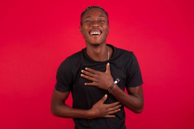 Neşeli Afrikalı Amerikalı erkek öğrenci ellerini çapraz yapar, güzel şakalara güler, günlük kıyafetler giyer, evde dikilir. Uzun saçlı mutlu genç adam duvara poz veriyor..