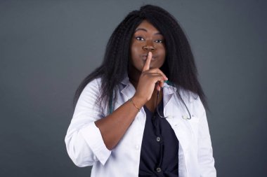 Sırrımı söyleme ya da yüksek sesle konuşma, lütfen! Duygusal olarak şaşırmış Afrikalı Amerikalı kadın doktor sessiz ol diyor, sessiz ol diyor, korkmuş bir ifadeyle sırrını açıklamaya korkuyor, gri stüdyo duvarına karşı duruyor..