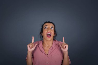 Esmer bir kadın, olumlu bir ifadeyle, iki işaret parmağıyla işaret ediyor, günlük kıyafetler giyiyor, geniş bir gülümsemesi var, gri arka planda izole edilmiş..