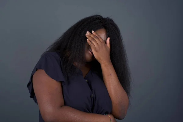 Indoor portrait of beautiful woman with stylish haircut, wearing casual clothes, making facepalm gesture while smiling, standing over gray background amazed with stupid situation.