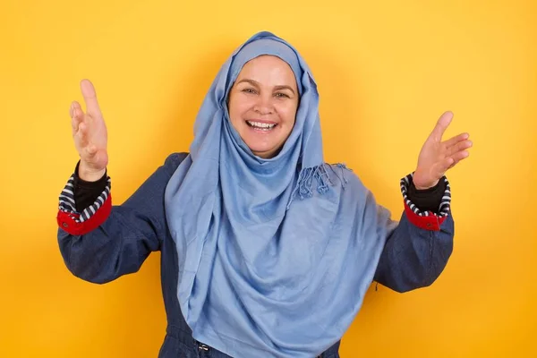 ヒジャーブで陽気な中年イスラム教徒の女性は 背景に隔離された頭の上に腕を上げる歓迎のジェスチャーを作ります — ストック写真