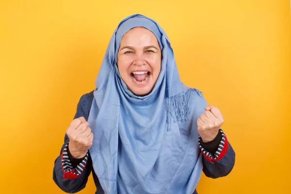 ヒジャブで中年のイスラム教徒女性の肖像画を閉じる 輝く背景の大きな目と広い開いた口に隔離された拳を上げる — ストック写真