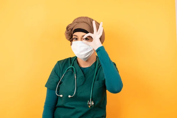 緑色の医療服の若い美しいイスラム教徒の医師の女性は 笑顔で指を通して見て手でOkのサインを行います 壁に立っている医療用ユニフォームを着用 — ストック写真