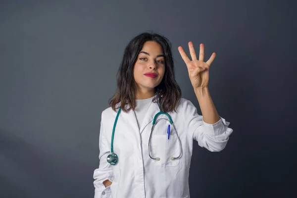 Young doctor woman standing against gray wall showing and pointing up with fingers number four while smiling confident and happy.