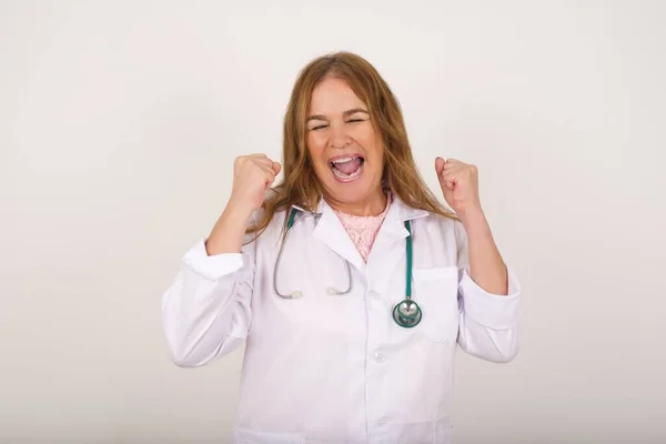 Beautiful young doctor woman wearing medical uniform being happy and excited expressing winning gesture. Successful and celebrating victory, triumphant, gray background.