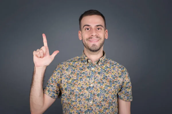 Kafkasyalı genç adam gri duvarın önünde duruyor. Bir numaralı parmaklarıyla işaret ediyor ve gülümsüyor. Kendinden emin ve mutlu..