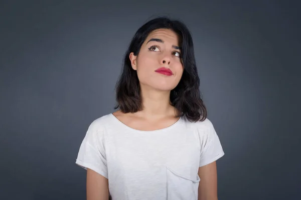 Troublesome puzzled Caucasian woman frowns face, bites lip, raises eyebrows, looks up, has problems, dressed in fashionable clothes, isolated over gray background, has regret look.