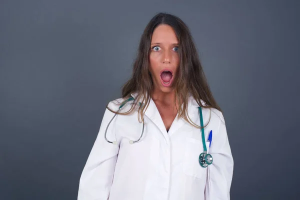 有表情的面部表情身穿医疗制服的惊慌失措的女医生 被她在空白灰墙上看到的东西惊呆了 — 图库照片
