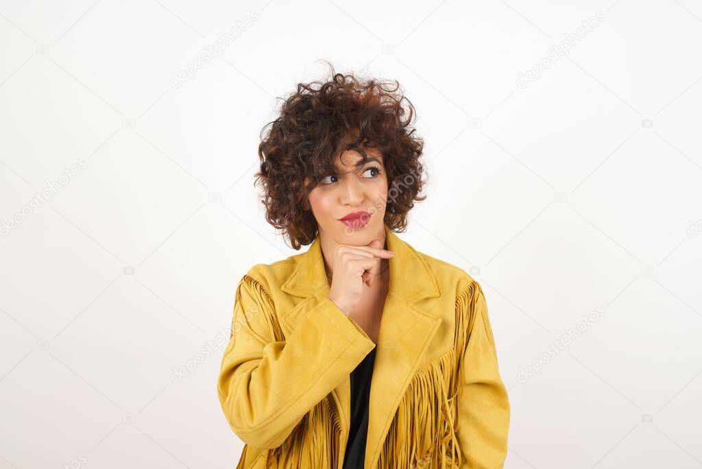 Dreamy   woman   with pleasant expression, wearing casual clothes, looks sideways, keeps hand under chin, thinks about something pleasant, poses against gray background.