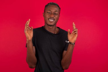 Kırmızı arka planda siyah tişört giyen yakışıklı Afro-Amerikan bir adam. Patronunun işyerinde yaptığı hataları fark etmemesini umarak parmaklarını çapraz yapıyor..