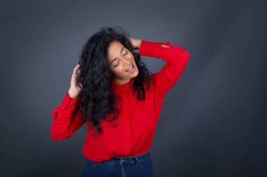 Kıvırcık saçlı, kırmızı tişörtlü, kendine güvenen bir ifadeyle gülümseyen ve ellerini başının üstünde tutan çekici esmer kadın portresi. Gri arkaplanda duruyor.