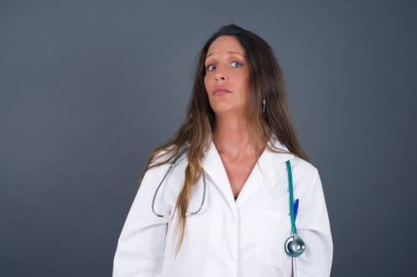 Züppe ifadeli güzel bir doktor kadın dudaklarını kıvırıyor ve kaşlarını kaldırıyor şüpheli ve şüpheci bir ifadeyle şüpheli ve şüpheci bir ifadeyle bakıyor. Gri arka planda kapalı duruyor..