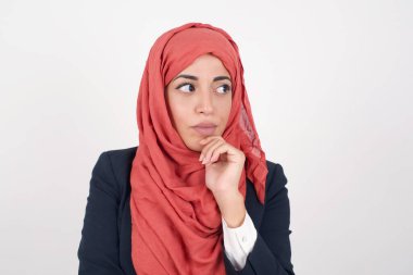 Düşünceli, düşünceli, güzel Müslüman kadın siyah ceket giyiyor ve tesettür elini çenenin altında tutuyor, düşünceli bir şekilde yukarı bakıyor, günlük kıyafetler giyiyor, beyaz arka planda mesajınız için boş bir alan bırakıyor.