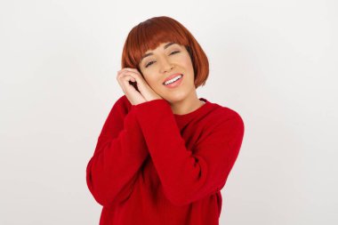 Tatlı yüzlü, hayalperest genç bir kadın, gözlerini kapatıyor, ellerini yüzüne çarpıyor, hoş bir şey düşünüyor, gri arka planda poz veriyor..