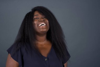 Güzel Afro-Amerikalı kadın, geniş gülümsemeli, beyaz dişli, sıradan tişörtlü, gri stüdyo duvarının üzerinde duran, izin gününün keyfini çıkarıyor. Kadın içeride duruyor..