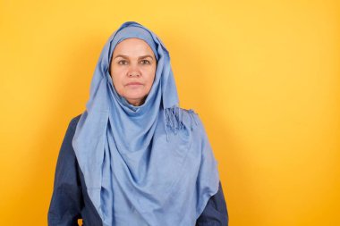 Orta yaşlı, neşeli, tesettürlü Müslüman bir kadının kameraya bakıp düşünmesi ya da merak etmesi. İki kolunu da indir, tarafsız yüz ifadesi..