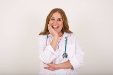  Tıbbi üniforma giyen yetişkin, beyaz bir kadın. İnanamamasına şaşırmış, ağzı açık, eli yüzüne yakın bir kadın. Olumlu duygu yüz ifadesi vücut dili.