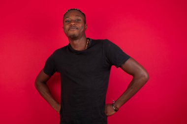 Kırmızı arka planda siyah tişört giyen komik, yakışıklı Afro-Amerikan bir adam. Şık bir saç kesimi ve sakalı var. El ele tutuşup tuhaf bir duruma bakıyor..