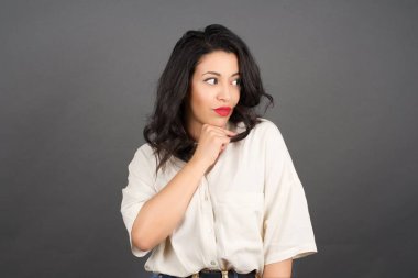 Güzel esmer kadın, sıradan giyinmiş, gözlerini kaçırıyor. Gri arkaplanda duruyor.  