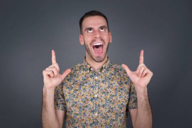 Olumlu ifadeli, kaygısız, her iki işaret parmağıyla işaret eden, günlük kıyafetler giymiş, geniş ilgi çekici bir gülümsemesi olan, gri arka planda izole edilmiş bir adam..