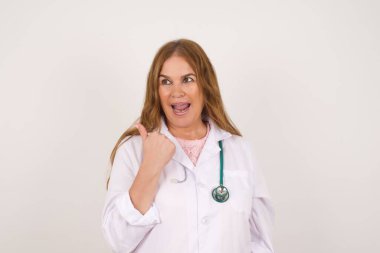 Omg konsepti. Şaşırmış bir şekilde tıbbi üniforma giyen, kafası karışmış, ağzı ve gözlerini açan, baş parmağıyla işaret eden, beyaz arka planda garip bir şey gösteren yetişkin, beyaz bir doktor kadın. Reklam kavramı.