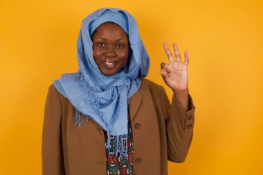 Çekici Afrikalı Amerikalı Müslüman bir kadının, olumlu bir ifadeyle, olumlu bir ifadeyle, olumlu bir işaret göstermesine sevindim. Sarı duvara yaslanarak.