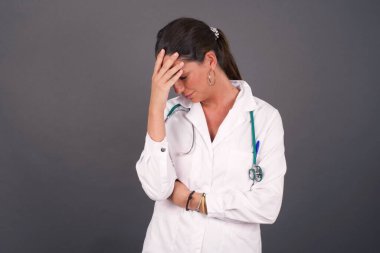 Güzel doktor kadının kapalı alanda portresi, tıbbi üniforma giymesi, gülümserken facepalm hareketi yapması, gri arka planda dikilmesi, aptalca durum karşısında hayrete düşmesi..