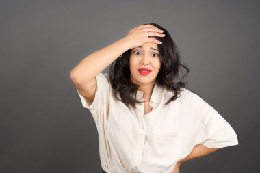 Gayri resmi giyinmiş güzel esmer kadının kafası karışmış. Gri arkaplanda duruyor. 