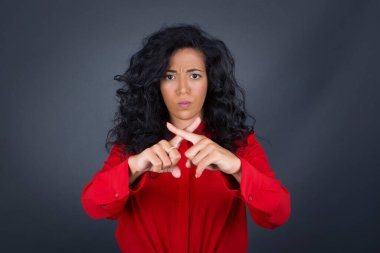 Kıvırcık saçlı, kırmızı tişörtlü, genç, güzel esmer kadın parmaklarını çapraz yapıp gri arka planda negatif işaret veriyor.