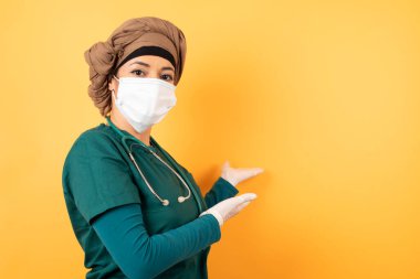 Yeşil üniformalı, genç, Müslüman doktor kadın izole edilmiş arka planda gülümseyerek içeri girmeye davet ediyor. Hoş geldin işareti.