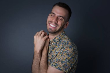 Hayalperest genç adam, hoş bir ifadeyle, gözlerini kapatır, ellerini yüzüne çapraz tutar, hoş bir şey düşünür, gri arka plana karşı poz verir..