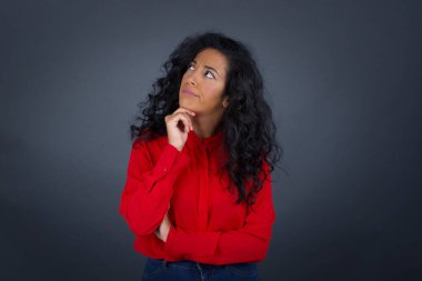 Kırmızı tişörtlü, kıvırcık saçlı, düşünceli esmer kadın portresi elini çenesinin altında tutuyor, yan bakıyor, ilginç bir şey düşünüyor ya da merak ediyor, sıradan giyinmiş, gri stüdyoya karşı poz veriyor..