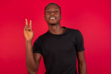 Genç Afro-Amerikan adam duvarın önünde duruyor ve iki numaralı parmaklarıyla işaret ediyor kendinden emin ve mutlu gülümserken..