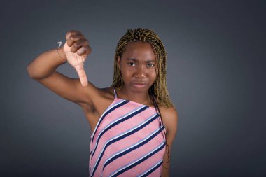 Hoşnutsuz genç Afro-Amerikalı kadın onaylamadığını gösteriyor, başparmaklarını aşağıda tutuyor, hoşnutsuzluğunu dile getiriyor, gri arka planda izole edilmiş hoşnutsuzlukla surat asıyor..
