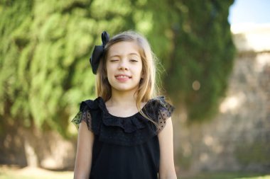 Gülümseyen ve dil gösteren mutlu ve komik yüzlü siyah elbiseli güzel bir genç kız. Günlük giysiler giyip parkta dikiliyor. 